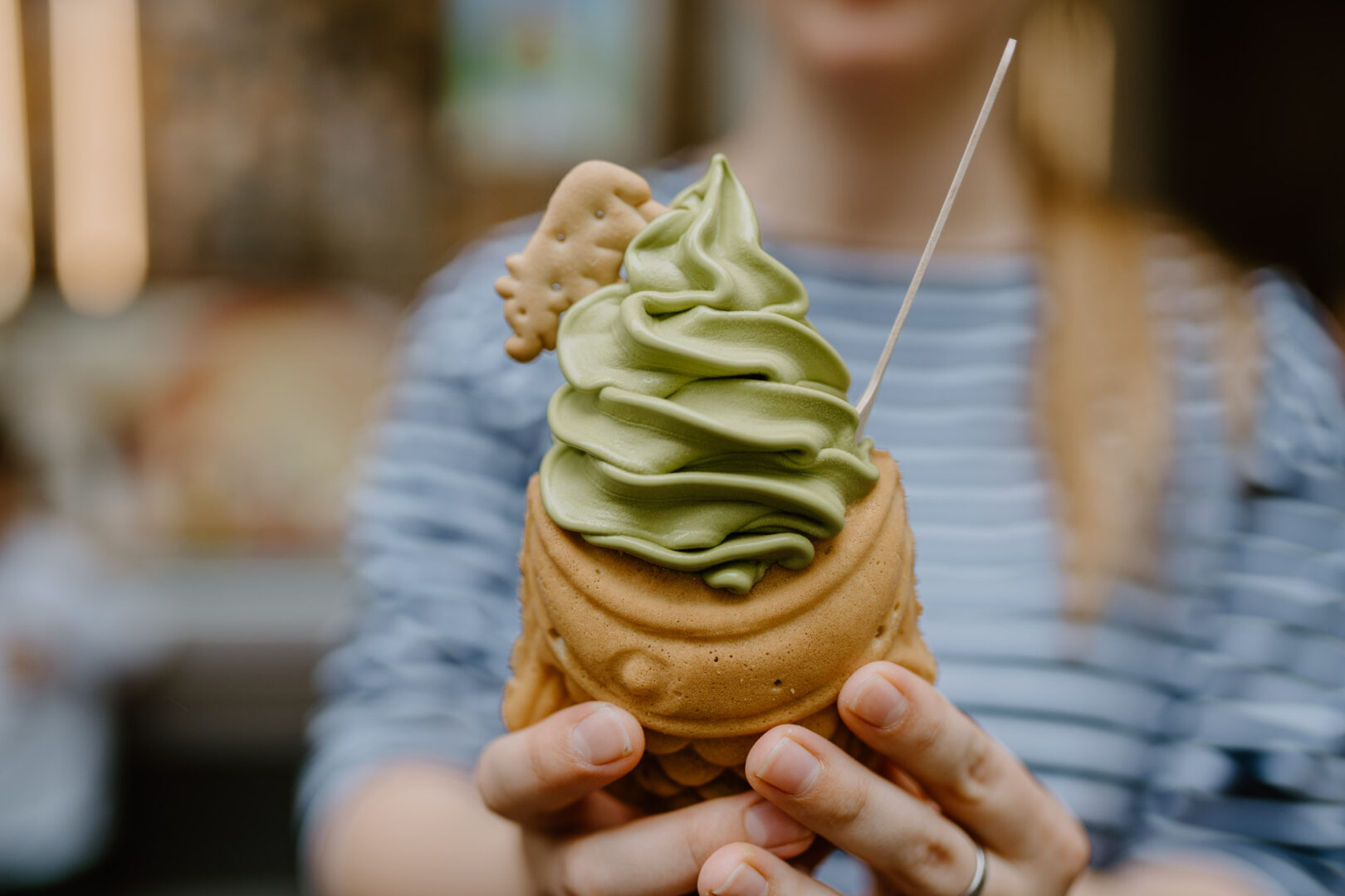 Man sieht ein Taiyaki, das japanische Gebäck in Fischform, gefüllt mit einer grünen Matchafüllung und in die Kamera gehalten von zwei Frauenhänden.