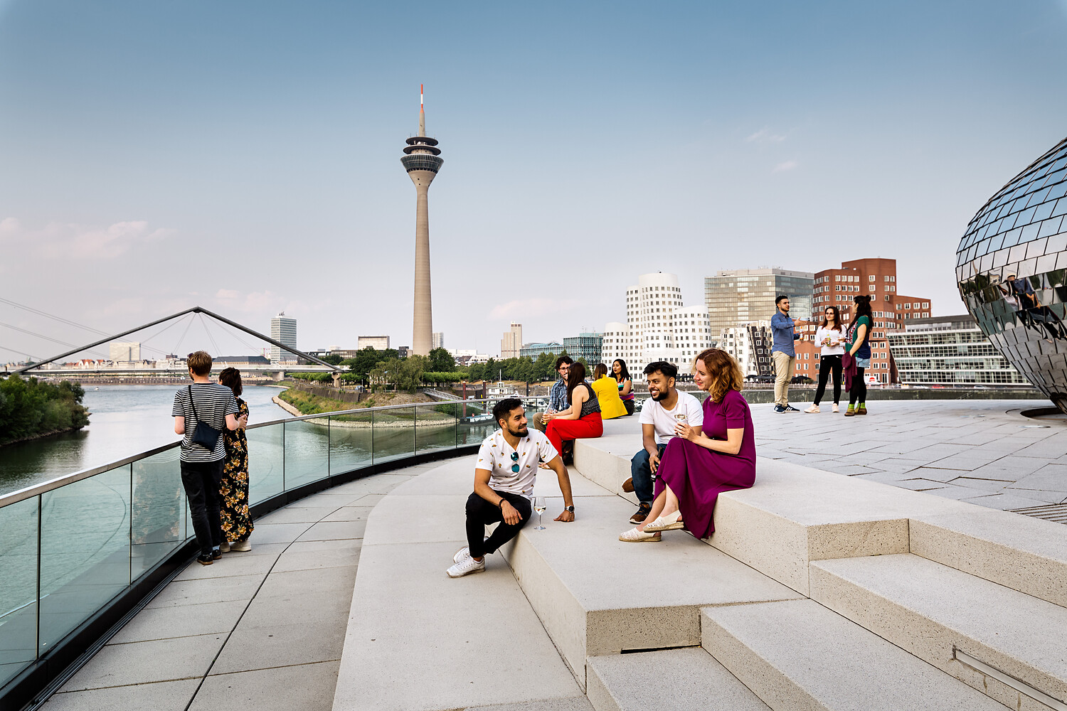 Man sieht eine Gruppe Menschen auf Stufen im MedienHafen, im Hintergrund der Rheinturm, die Gehry-Häuser.