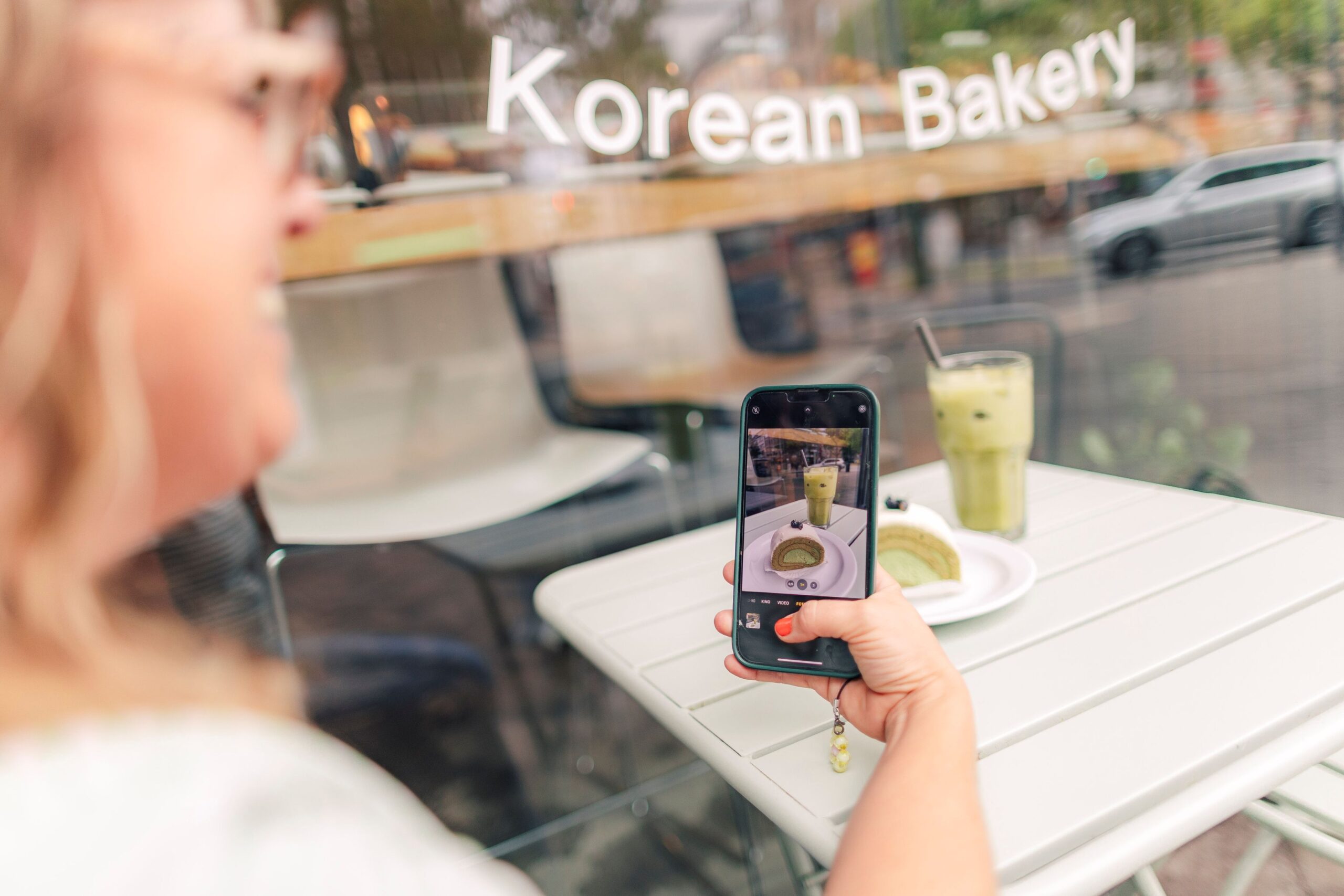 Man sieht links im Anschnitt eine Frau unscharf, sie hält ein Hancy in der Hand und fotografiert ihren Matcha-Cake und ihre Iced Matcha Latte.