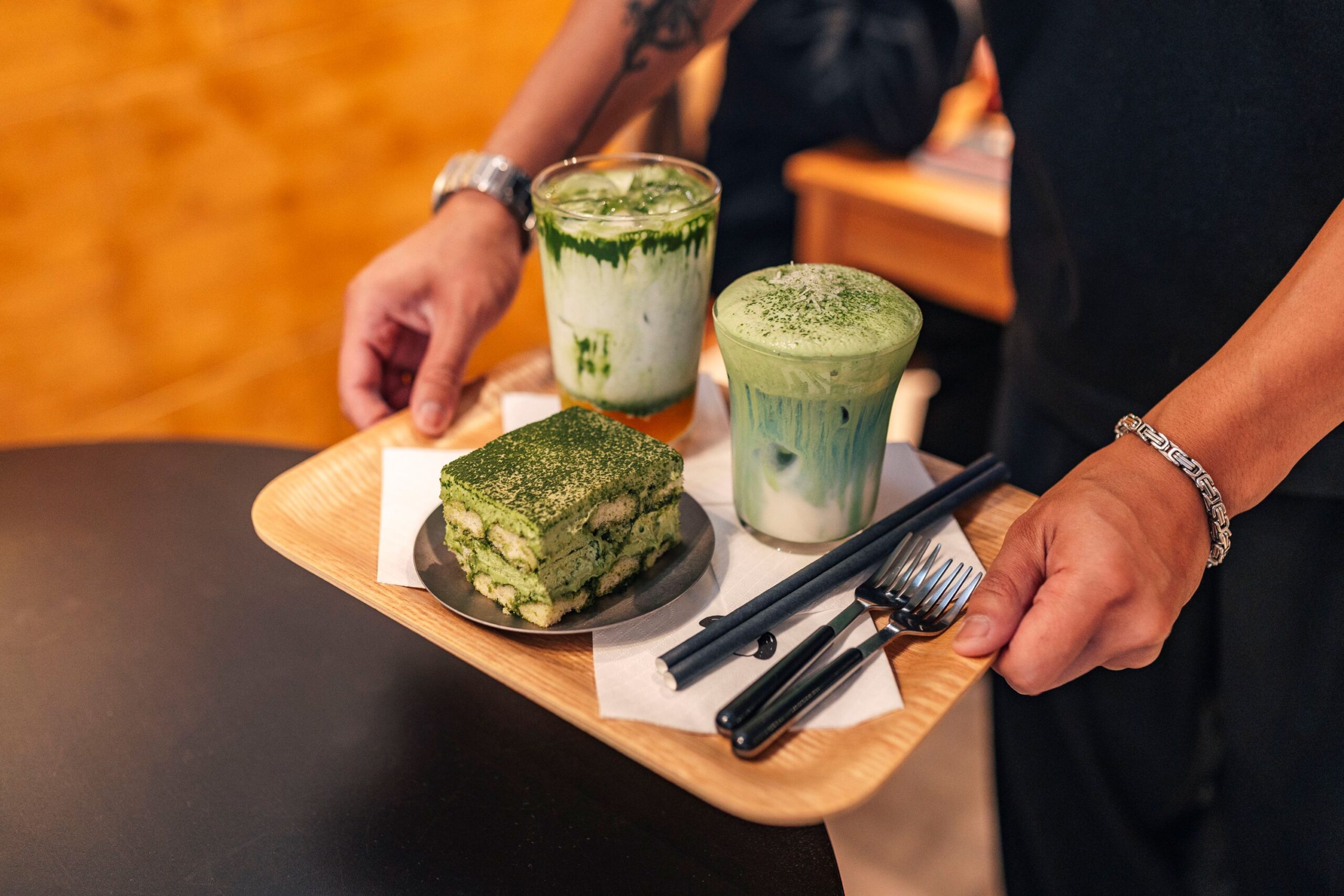 Man sieht Hände, die ein Tablett halten. Auf dem Tablett sind 2 Matcha-Getränke und ein Matcha-Tiramisu.