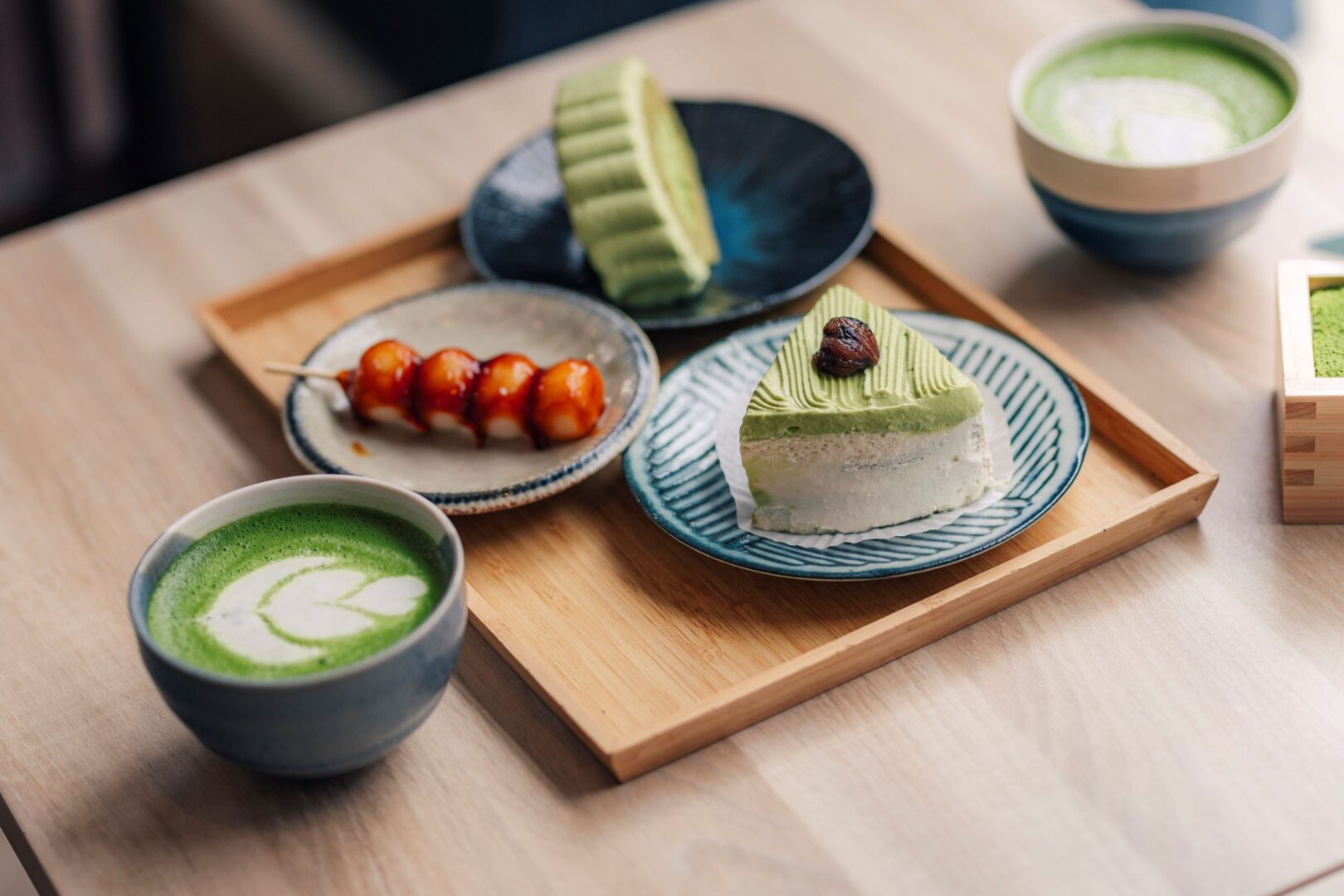 Man sieht ein Tablett auf einem Tisch mit einem Matcha in einer Keramikachale und drei Matcha-Desserts wie Kuchen.