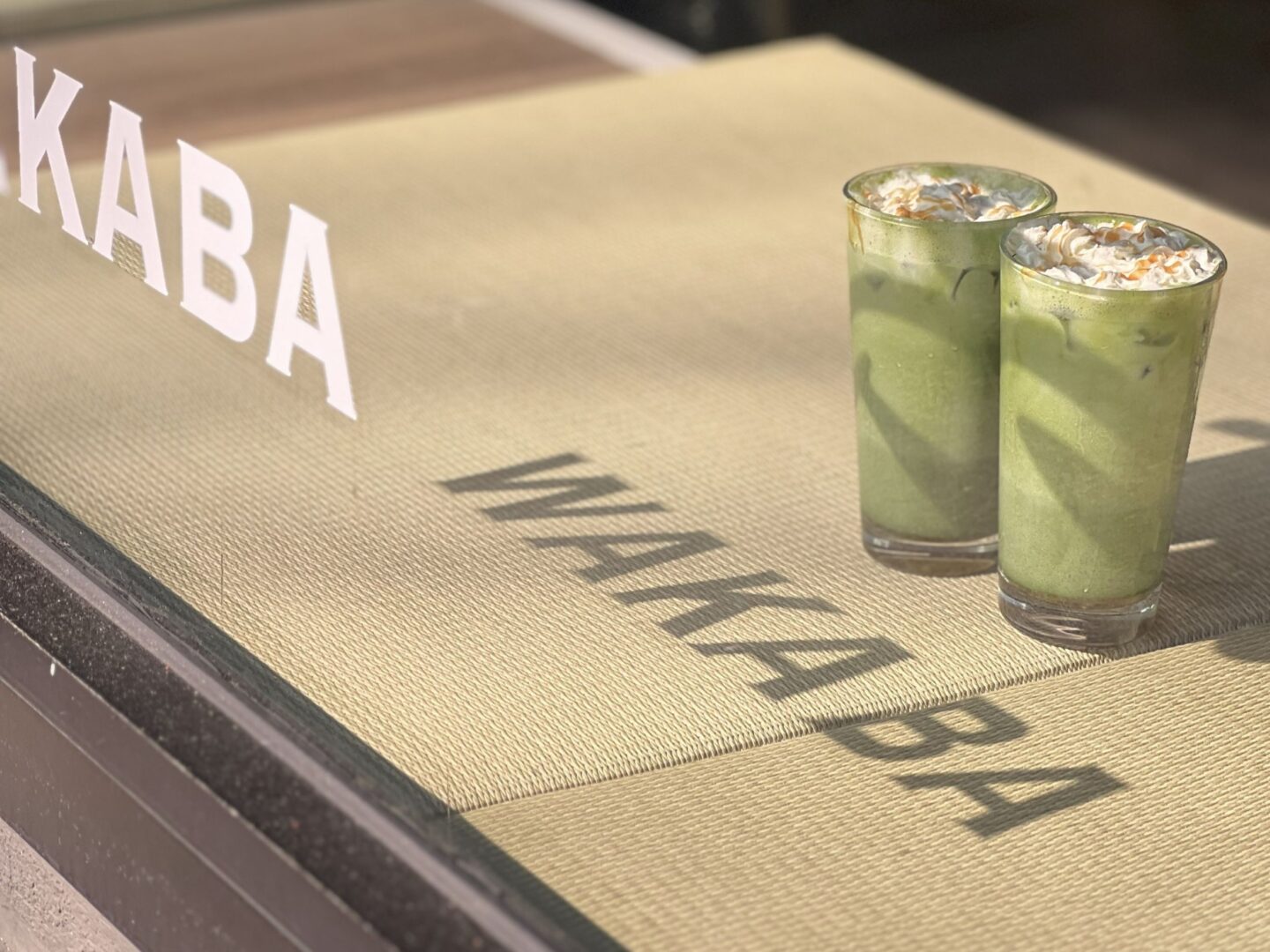 Man sieht durch das Schaufenster auf eine Ablage, auf der zwei Matcha Latte stehen und darunter als Schatten den Namen des Cafés: Wakaba.