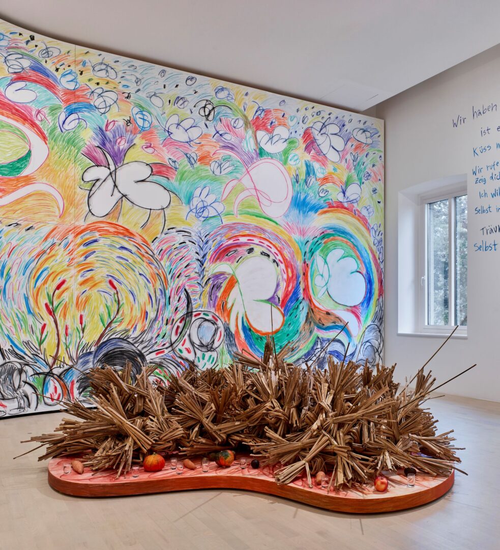 Ausstellungsansicht im K21: Man sieht ein wandfüllendes Gemälde, sehr farbenfroh, tendenziell blumig. Davor eine Installation, die etwas Wald-artiges hat.
