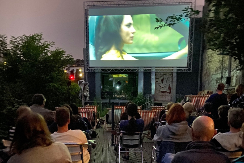 Man sieht eine Open-Air-Kinoveranstaltung. Die Leinwand mit einer Szene, auf der eine Frau im Auto sitzt und fährt, davor das Publikum auf Klappstühlen.