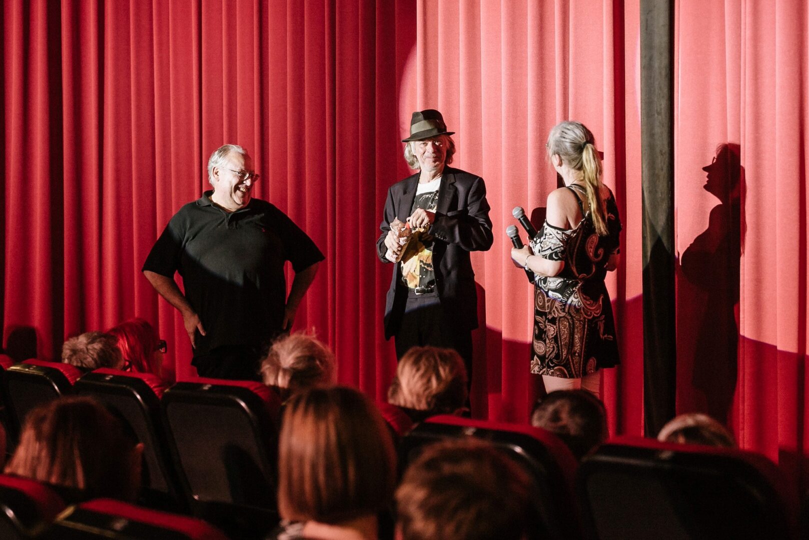 Man sieht Helge Schneider mit zwei weiteren Personen im Gespräch vor dem roten Vorhang im Cinema Kino.