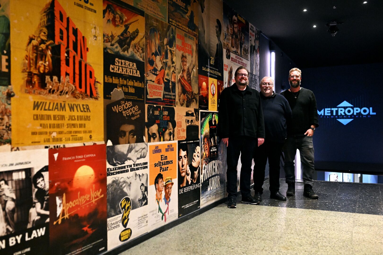Man sieht links eine Wand voller Filmplakate, am Ende stehen drei Männer vom Metropol Programmkino, hinter ihnen führt eine Treppe runter, darüber ein blaues Leuchtschild auf dem Metropol steht.