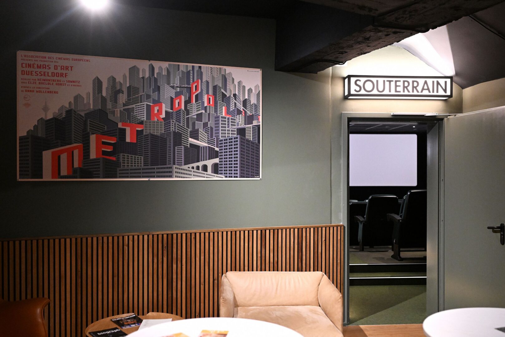 Man sieht rechts das Souterrain-Schild zu kleinen Kinosaal, links eine grüne Wand mit Plakat und Sesseln.