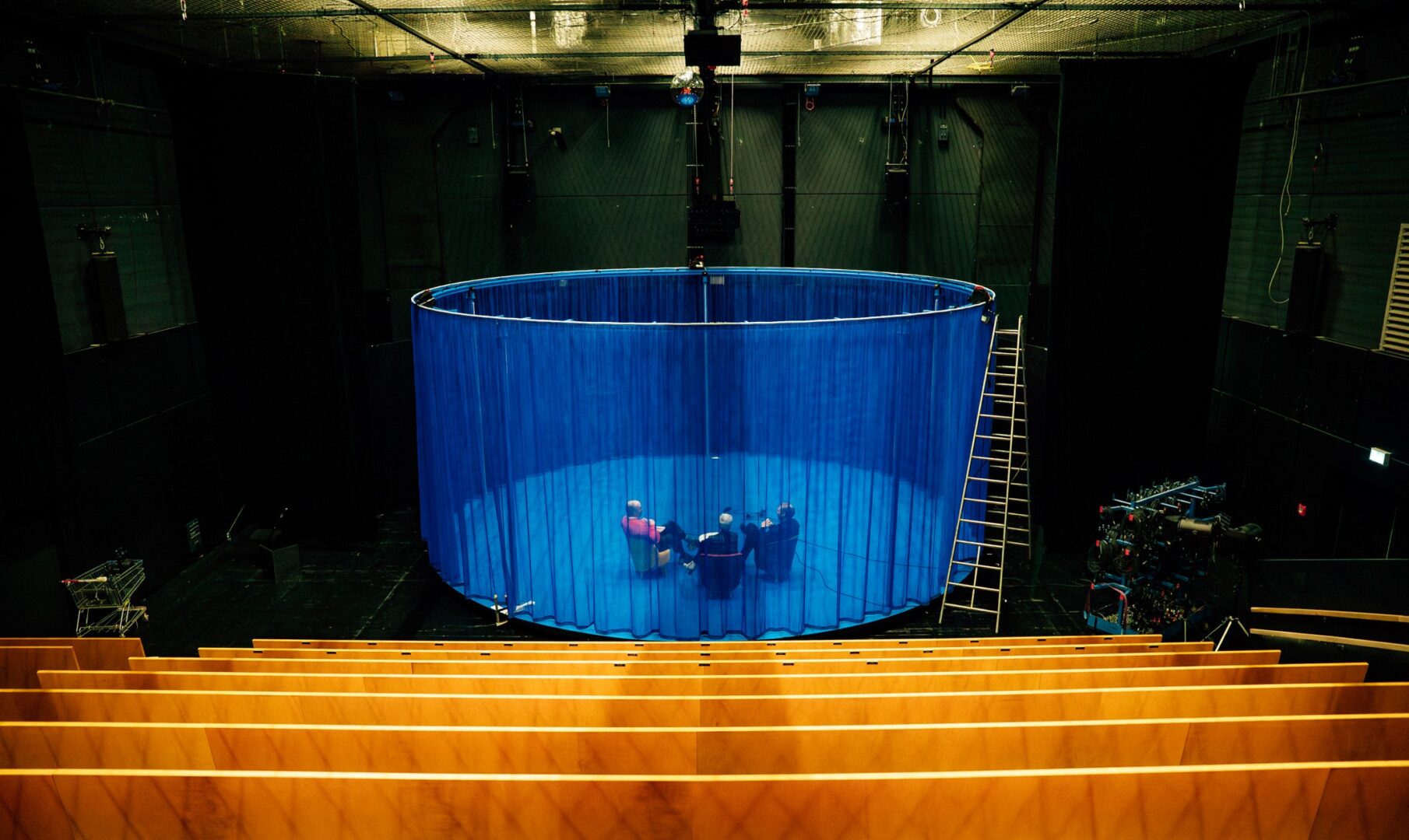 Man sieht vorne Bankreichen im Theater Central vom Düsseldorfer Schauspielhaus Im hinteren Drittel des Bildes die Bühne, in der Mittel der Bühne einen kreisförmig angelegten blauen Vorhang durch den man drei Personen sieht.