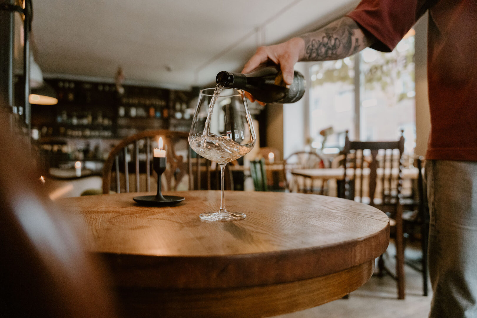 Man sieht wie jemand mit einem tätowierten Unterarm Weißwein in ein elegantes Weinglas einschenkt. Der Raum der WEinbar ist gemütlich mit viel Holz eingerichtet, ein bisschen Shabby-Chic-orientiert.