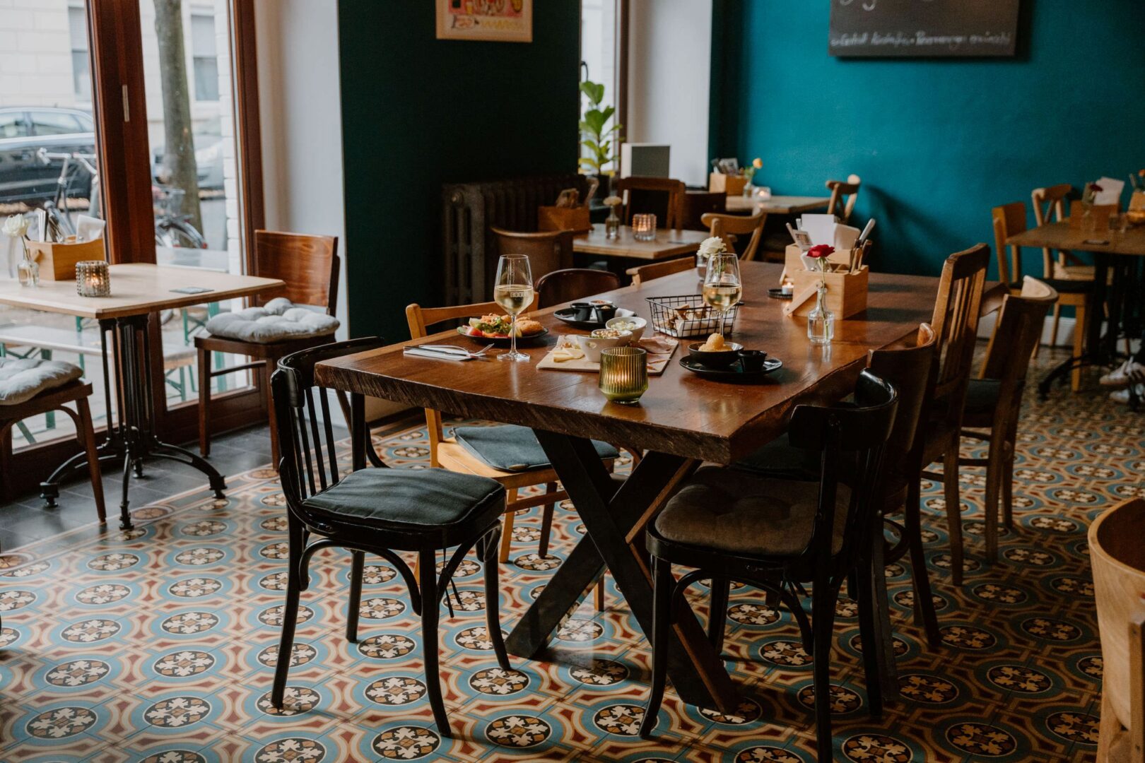 Cuvée ist eine Weinbar in Düsseldorf. Man sieht den Gastraum mit einem bunten Fliesenboden, petrolfarbenen Wänden und gemütlichen Holzstühlen an Holztischen.