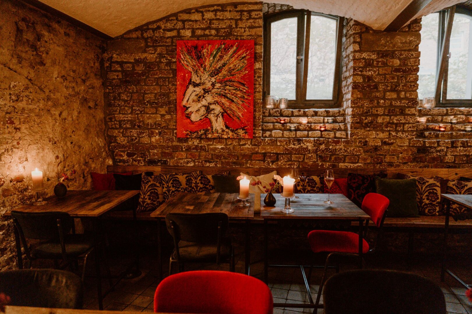Man sieht ein Kellergewölbe, das eine Weinbar in Düsseldorf ist. Auf den unverputzten Backsteinwänden hängt ein Bild, kleine Fenster lassen Licht rein. rustikale Holztische und rot gepolsterte Stühle wirken gemütlich.