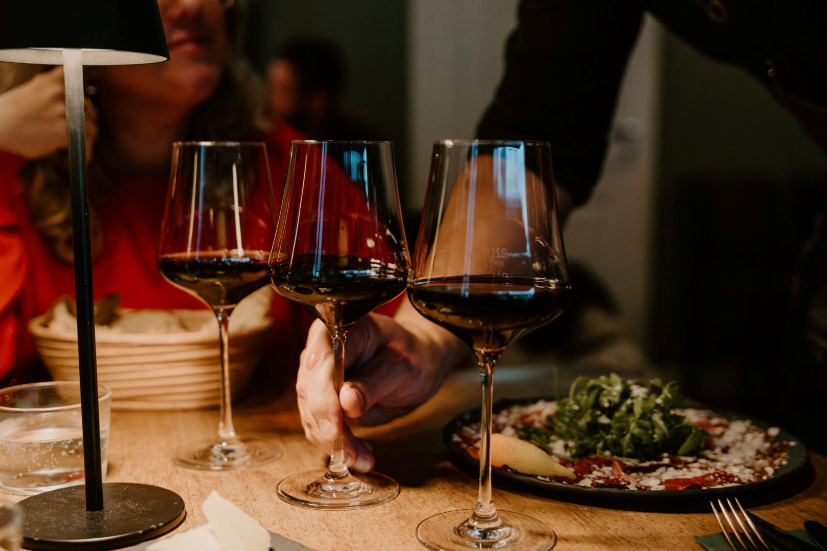 Man sieht drei Weingläser gefüllt mit Rotwein, eines wird von einer Hand gerade auf den Tisch gestellt, auf dem kleine Gerichte wie Carpaccio mit Ruccola stehen. Licht gedämpft und warm.
