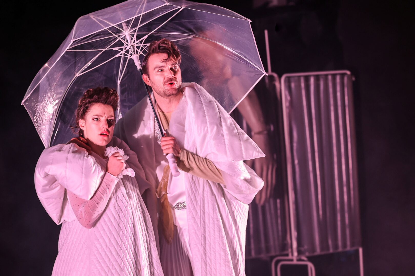 Szene aus Die Gischt der Tage: Sophie Stockinger mit einem Schauspielkollegen in weiß gekleidet, pink angeleuchtet unter einem transparenten Regenschirm.