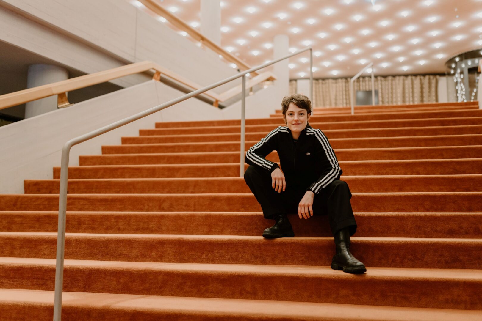 Sophie Stockinger sitzt auf der großen Treppe im Foyer des Düsseldorfer Schauspielhauses. Die Treppe ist mit dunkelorangem Teppich bezogen.