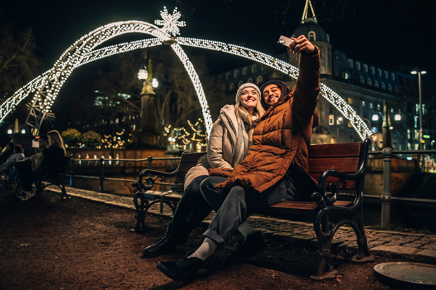 Ein Parchen sitzt auf einer Bank auf der Giradet-Brücke, die über den Kö-Graben verläuft. Das Pärchen macht ein Selfie mit dem Lichterdom im Hintergrund,.
