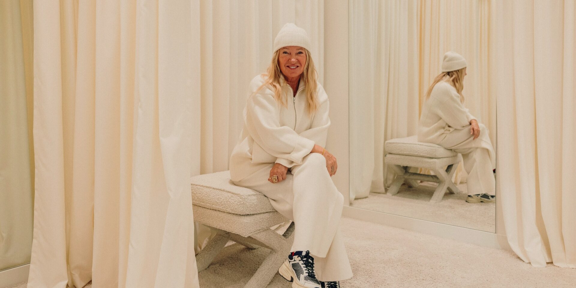 Evelyn Hammerström, Gründerin und Inhaberin des Concept Stores Jades, sitzt auf einem cremeweißen Hocker, in einem cremeweißen Outfit in einer Umkleide, deren Vorhänge und Teppich ebenfalls cremeweis sind.