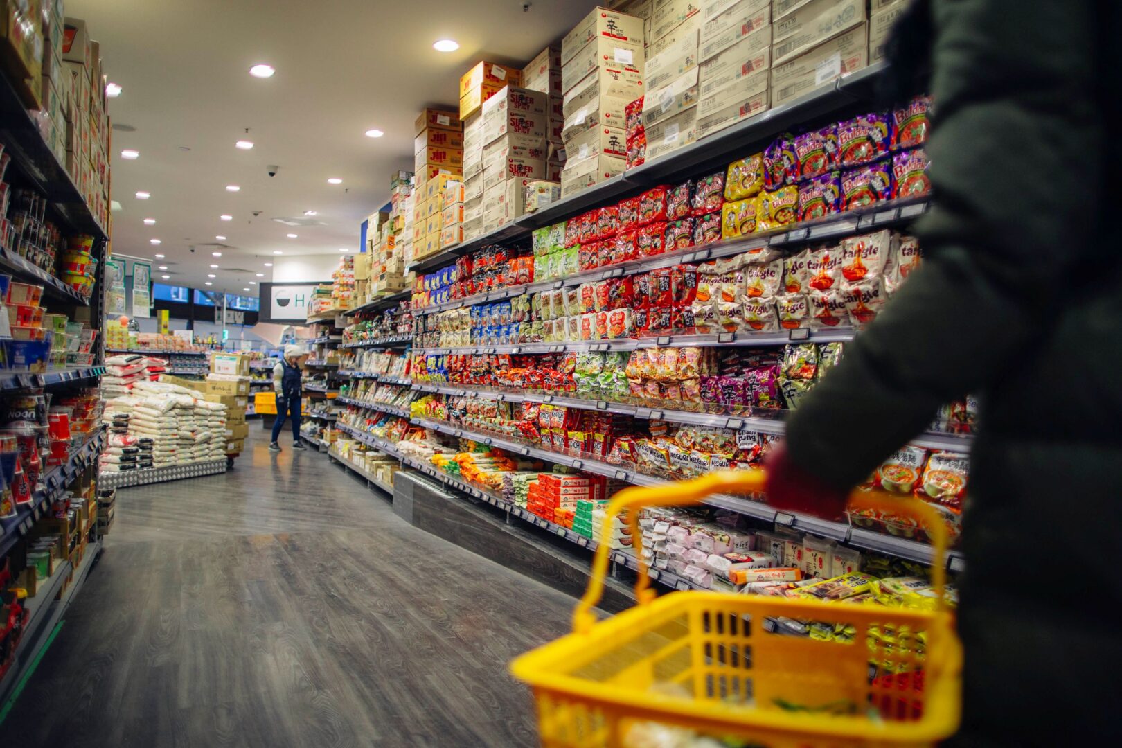 Man sieht einen langen Gang im asiatischen Supermarkt, vorne rechts angeschnitten einen Arm, dessen Hand einen gelborangenen Supermarkteinkaufskorb hält.