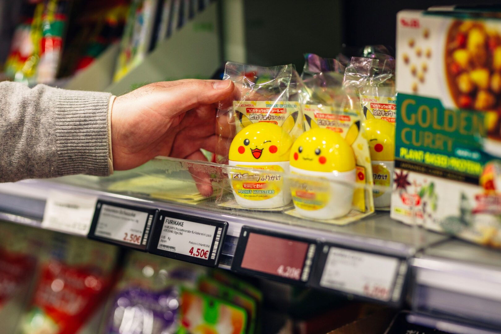 Eine Hand greift in ein Supermarktregal und holt eine Gewürzmischung heraus, die in einer niedlichen Box verpackt ist. Die Box sieht aus wie eine Comic-haftes Küken, ist gelb und ein Comic-Gesicht ist darauf gedruckt.