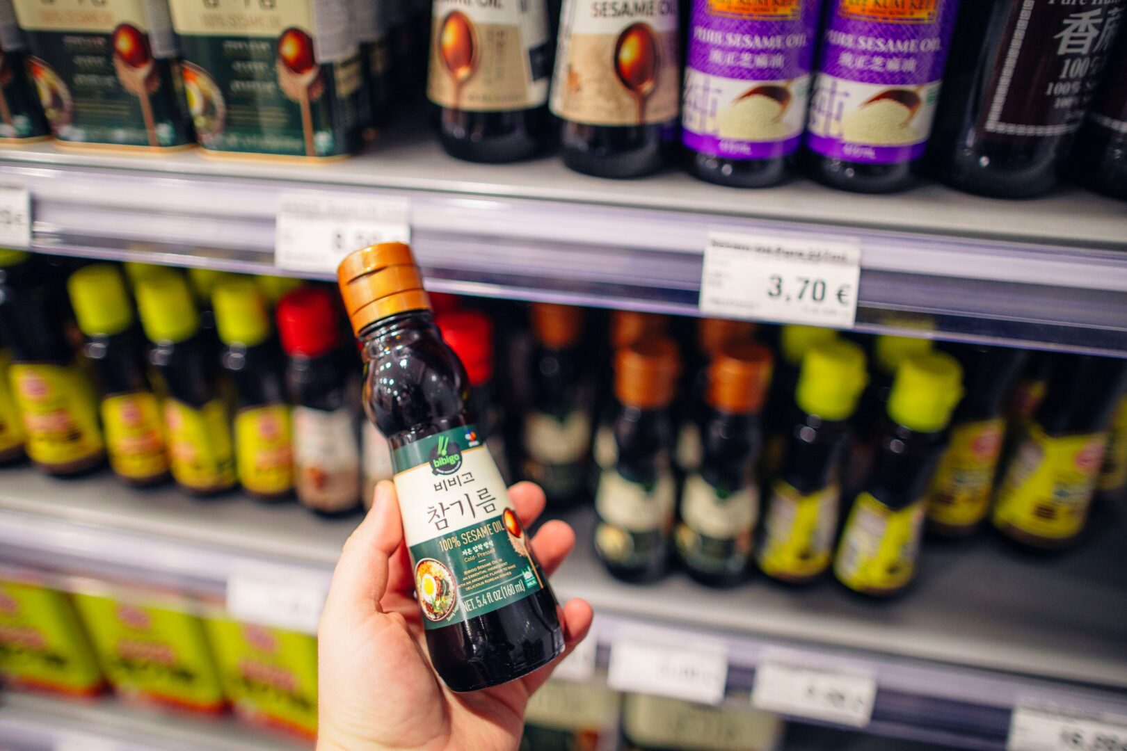 Eine Hand hält eine kleine Flasche Sesamöl, dahinter sieht man ein volles Supermarktregal.