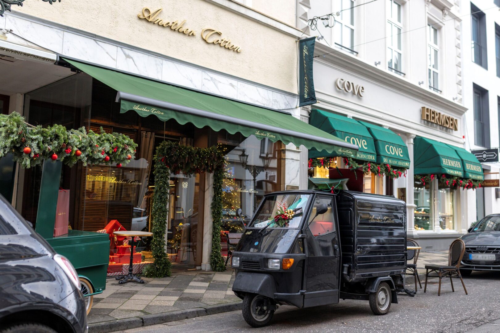 Man sieht eine kleine Straße mit Geschäften. In der Mitte Another Cotton Club mit grüner Markise und weihnachtlichem Tannengrün im Anschnitt.