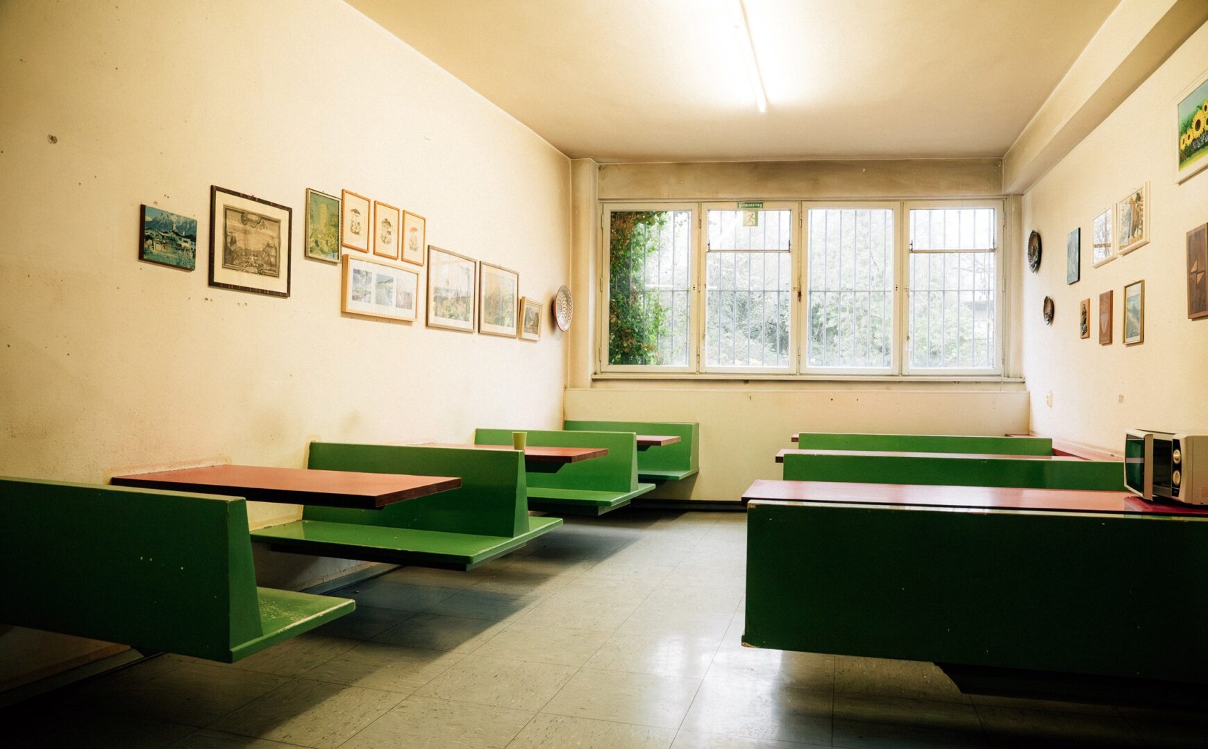 Man sieht in die menschenleere Kantine, rechts und links grüne Bänke und Holztische, an den Wänden hängen Fotografien.