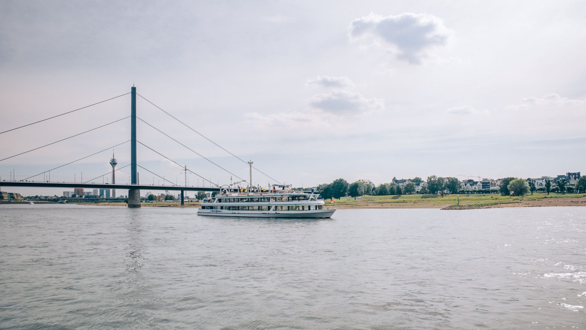 Ausblick auf den Rhein, man sieht im Hintergrund eine Brücke und den Rheinturm. Es ein Schiff ist auf dem Rhein zu sehen.