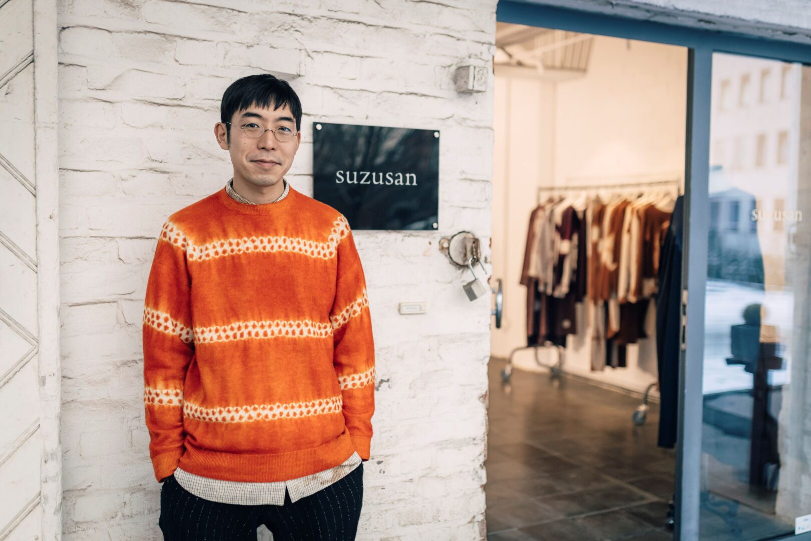 Hiroyuki Murase steht am Eingang vor dem Atelier seines Labels Suzusan. Er trägt einen orangefarbenen Pulli, der in einer Art Batiktechnik gefärbt wurde. 
