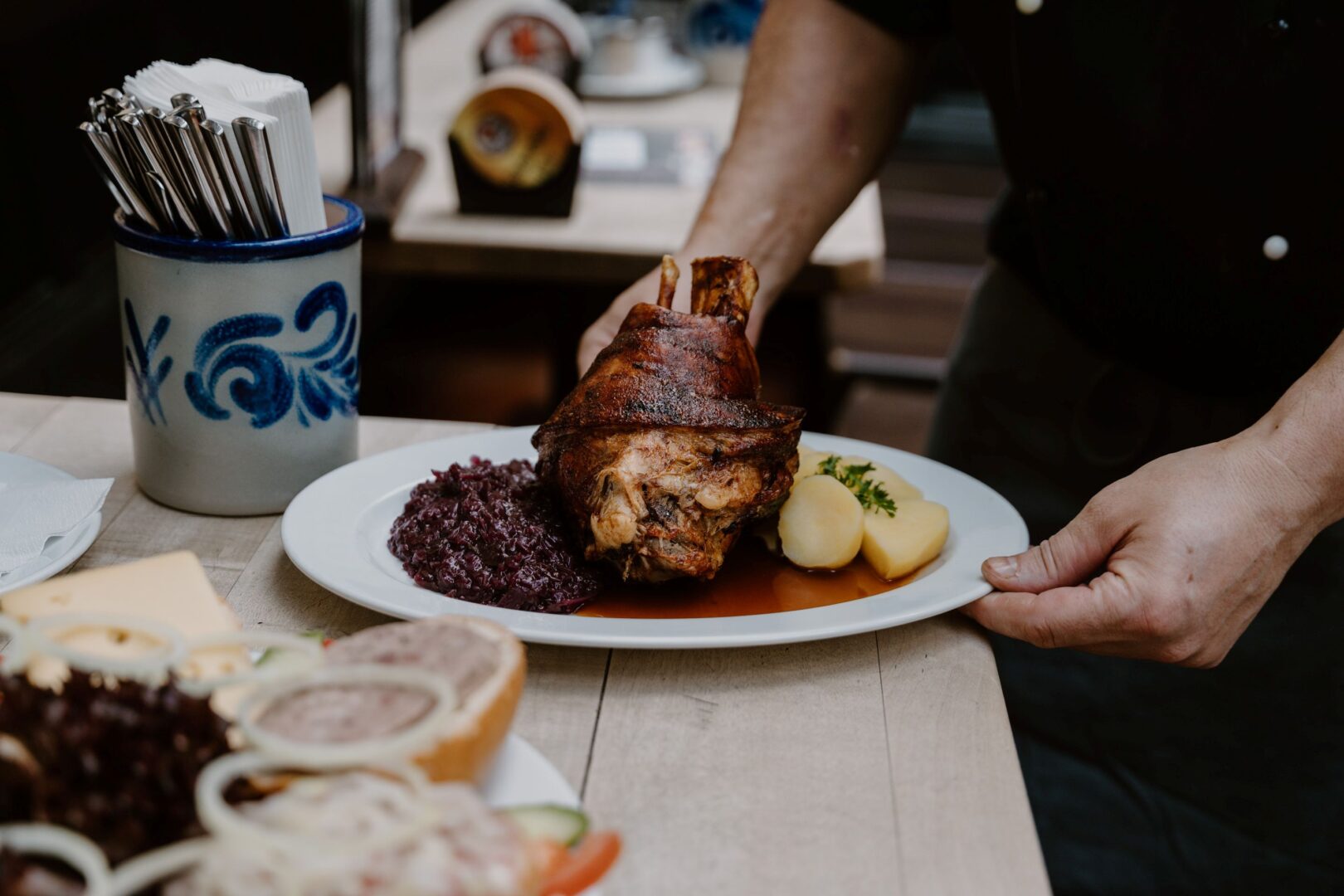 Man sieht die Hände eines Kellners, der einen Teller mit einer Haxe mit Salzkartoffeln und Rotkohl auf einen Tisch im Gastraum stellt. Es ist das Brauhaus Düsseldorf Im Füchschen.