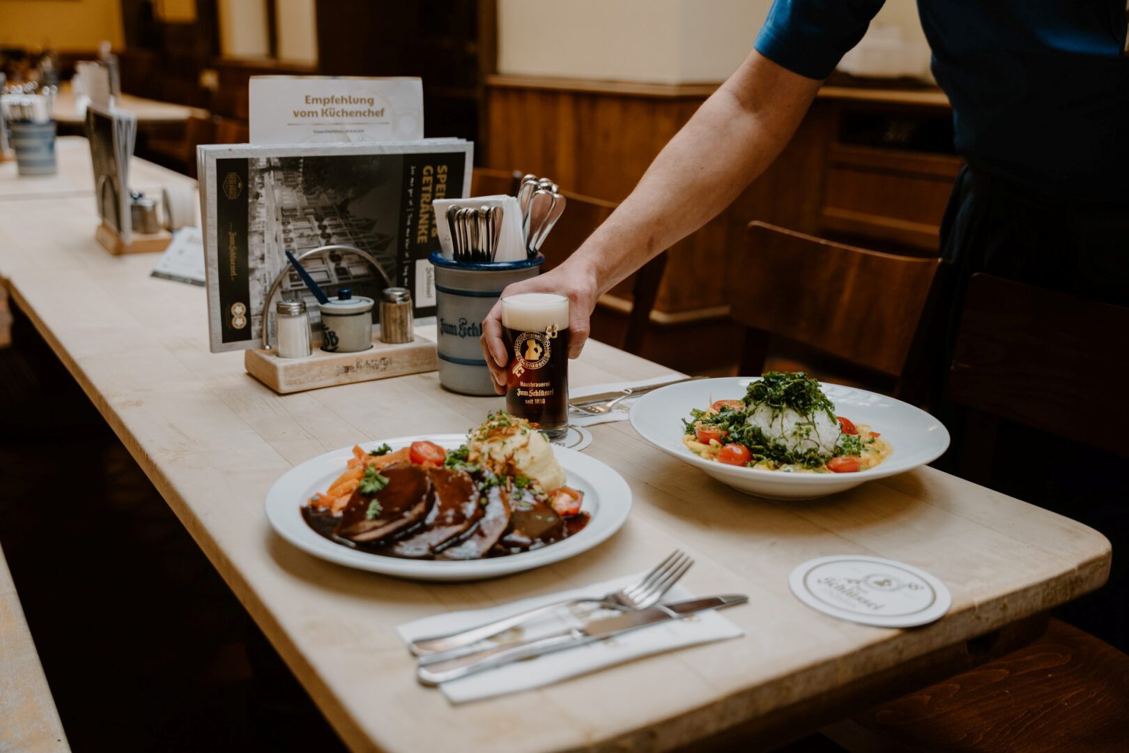 Im Brauhaus Düsseldorf Zum Schlüssel: Man sieht einen gedeckten Holztisch mit Sauerbraten und einem vegetarischen Salat und den Kellner im Anschnitt, der ein Glas Altbier dazustellt. 