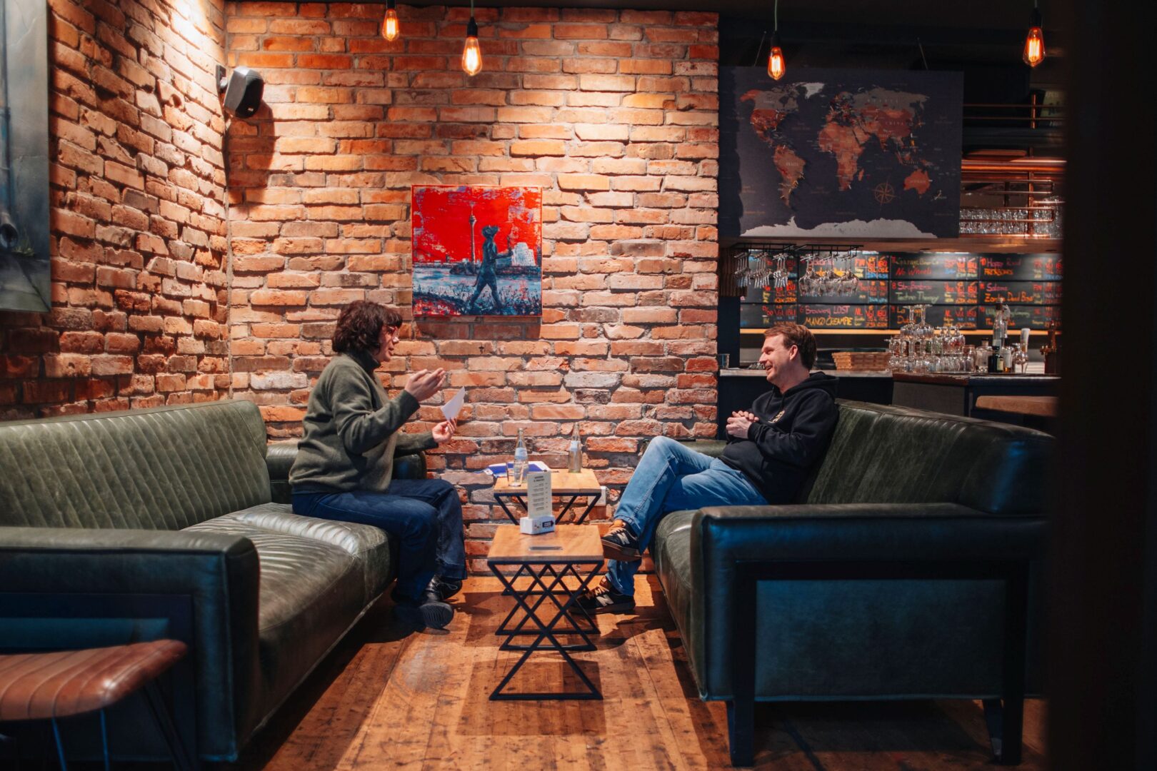 Katja (li) und Sebastian sitzen im Lokal vor einer Backsteinwand und reden miteinander. 