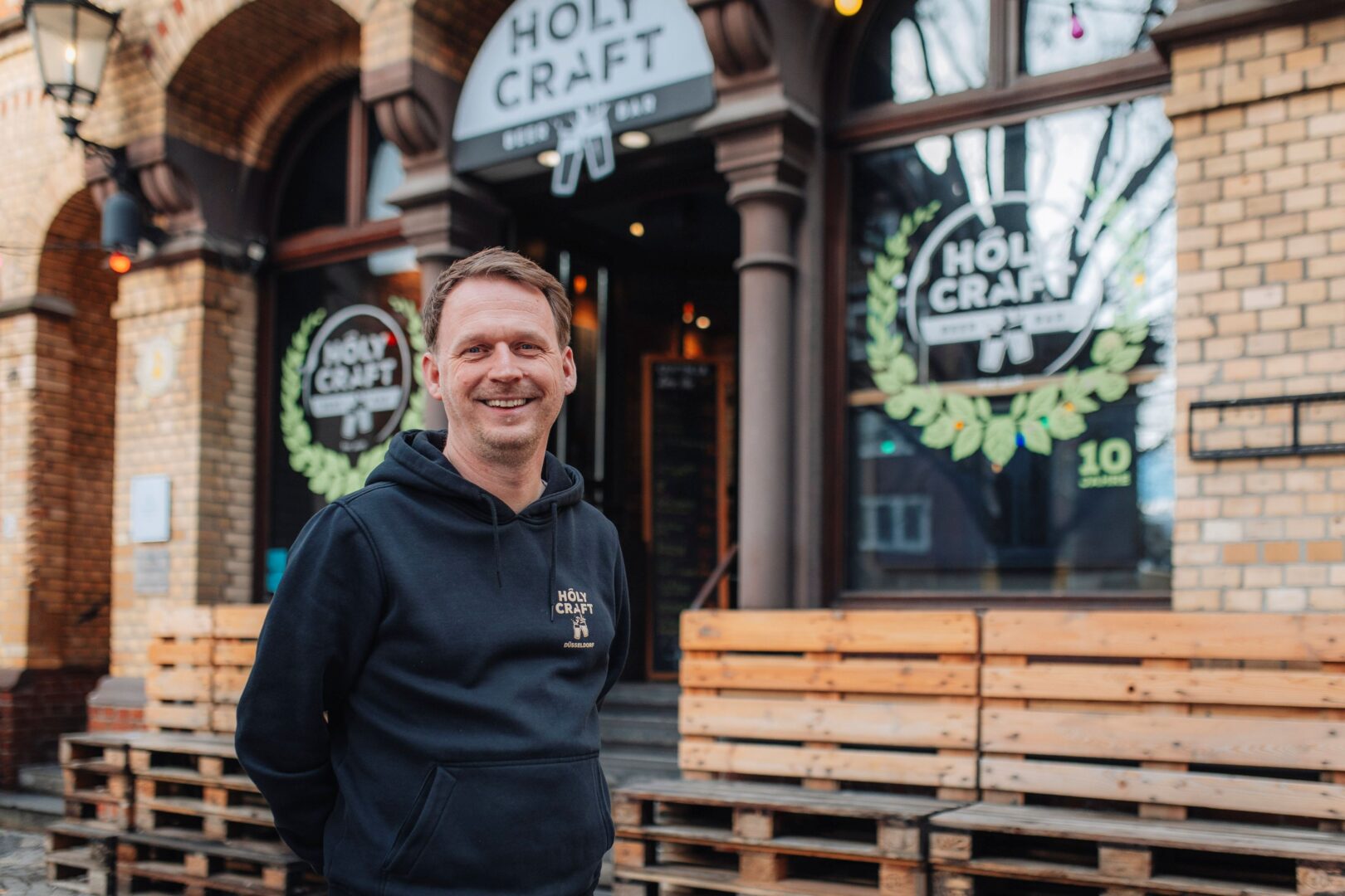 Sebastian trägt einen schwarzen Hoodie und steht vor der Holy Craft Beer Bar. Man sieht Holzbänke und Fenster, die mit dem Holy Craft Beer-Logo beklebt sind. 