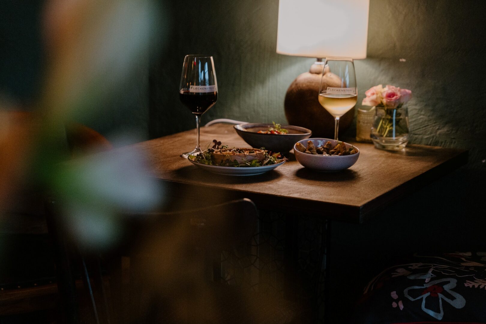 Das Wine and Dine-Lokal Fleckensteins. Man sieht durch eine unscharfe Pflanze links auf einen gedeckten Tisch mit Essen und 2 Gläsern Wein. 