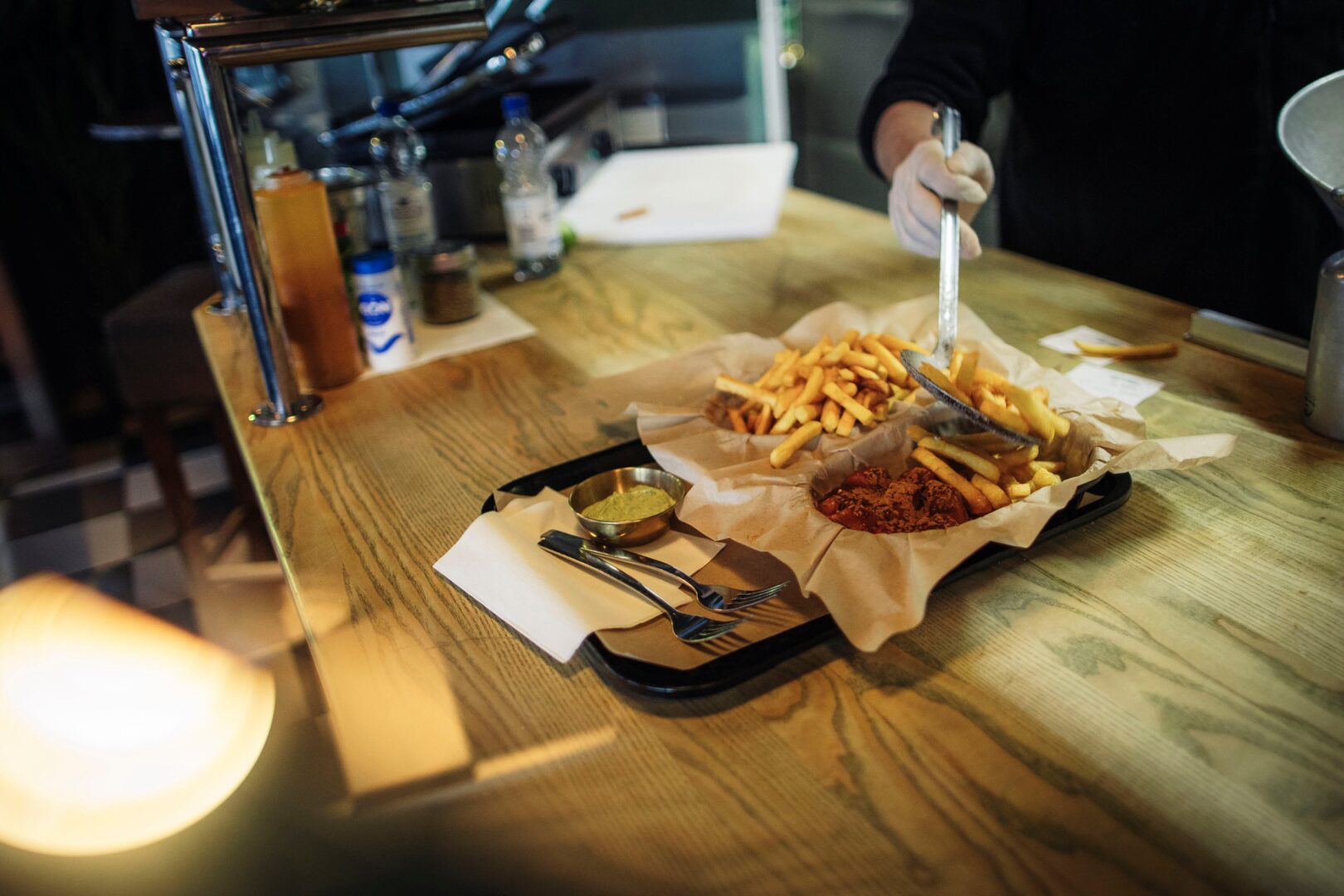 Man sieht ein Tablett, darauf zwei Schalen, die mit Backpapier-artigem Papier ausgeschlagen sind. Darin Pommes und Currywurst. 