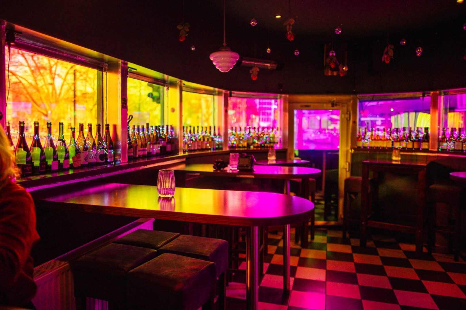 Man sieht einen Teil des Lokals Pommes und Wein: Das einstöckige Flachdachgebäude ist an den Seiten rund. Man sie die mit farbiger Folie abgeklebten Fenster im runden Teil des Restaurants, wodurch das Licht gelb und pink in den Raum mit schwarz-weiß gekacheltem Boden strahlt. 