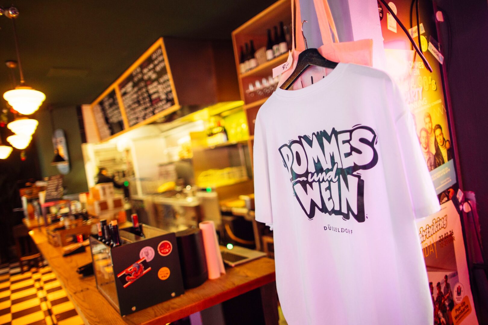 Man sieht im Lokal hängend ein weißes T-Shirt mit einem nach Graffiti aussehenden Aufdruck: Pommes und Wein. 