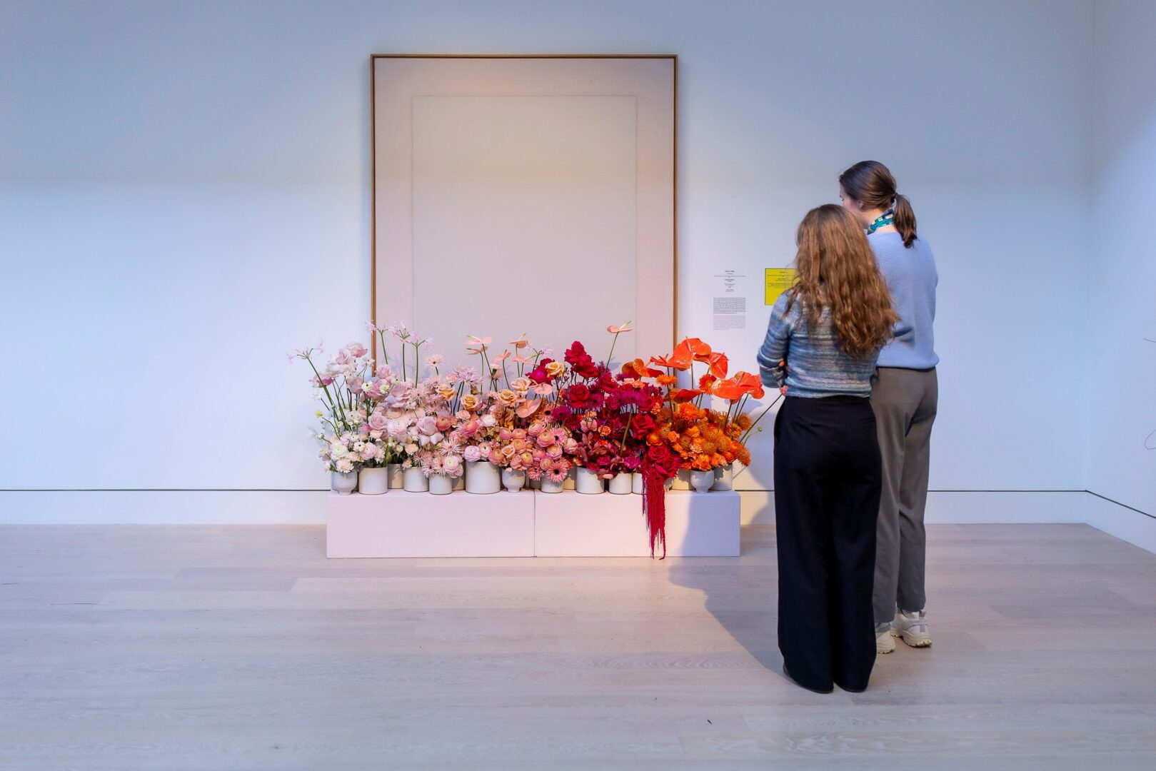 Ein Blumenarrangement von Victor Breuer im Rahmen der Ausstellung "Palastblühen" im Kunstpalast. Das Arrangement hat einen Farbverlauf von links nach rechts: weiß, rose, pink, rot nach orange. Es ist in einem weißen, rechteckigen Kasten, der genau abgemessen, so lang wie das abstrakte Gemälde von Ulrich Erben.