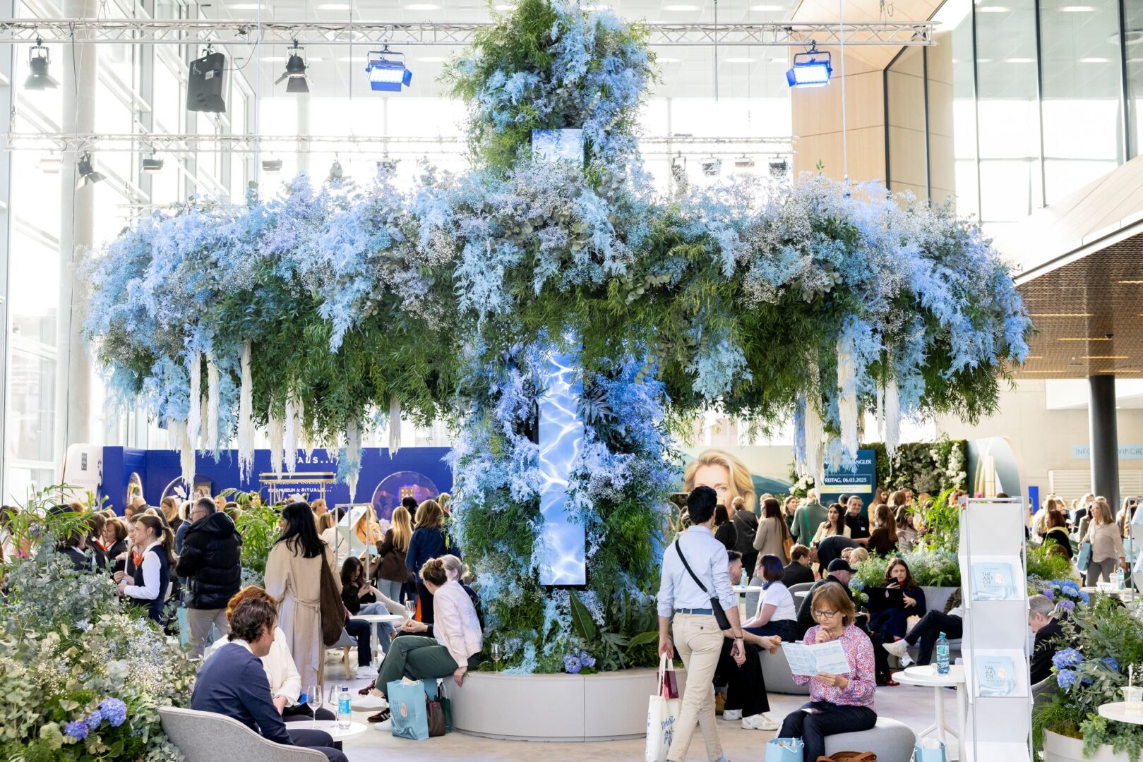 Man sieht eine Eingangshalle der Messe Düsseldorf während der B2C-Messe "The Art of Beauty & Health" im Zentrum ist ein raumfüllendes Blumenarrangement aus Blumen in verschiedenen Blauschattierungen, dass von der Decke wie ein überdimensionierter Blumenkranz herunterhängt. Darunter sitzen Besucher*innen der Messe.
