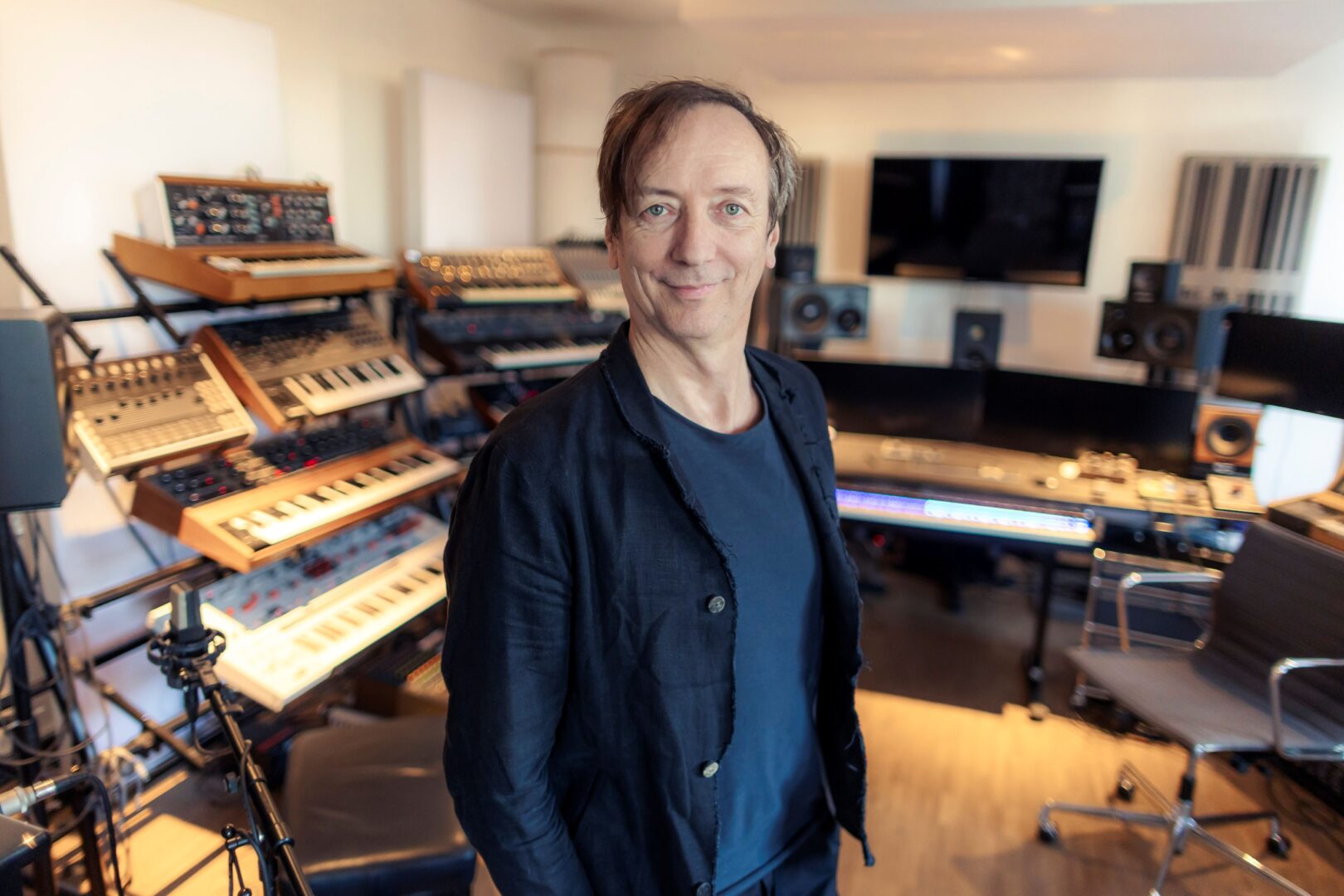 Volker Bertelmann steht im Studio, im Hintergrund sieht man Keybords und weitere Tasteninstrumente. 