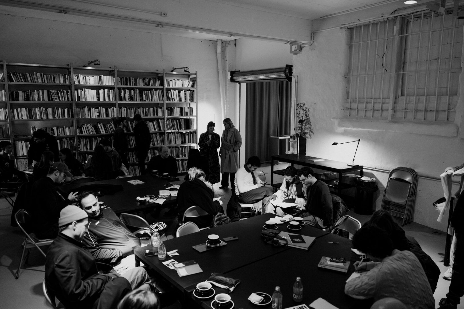 Schwarzweiß Foto eines Off-Spaces der Düsseldorfer Kunstszene. Man sieht einen großen Raum, im Hintergrund ein hohes Regal voller Bücher, davor Menschen die lesen, Musik hören und sich unterhalten.