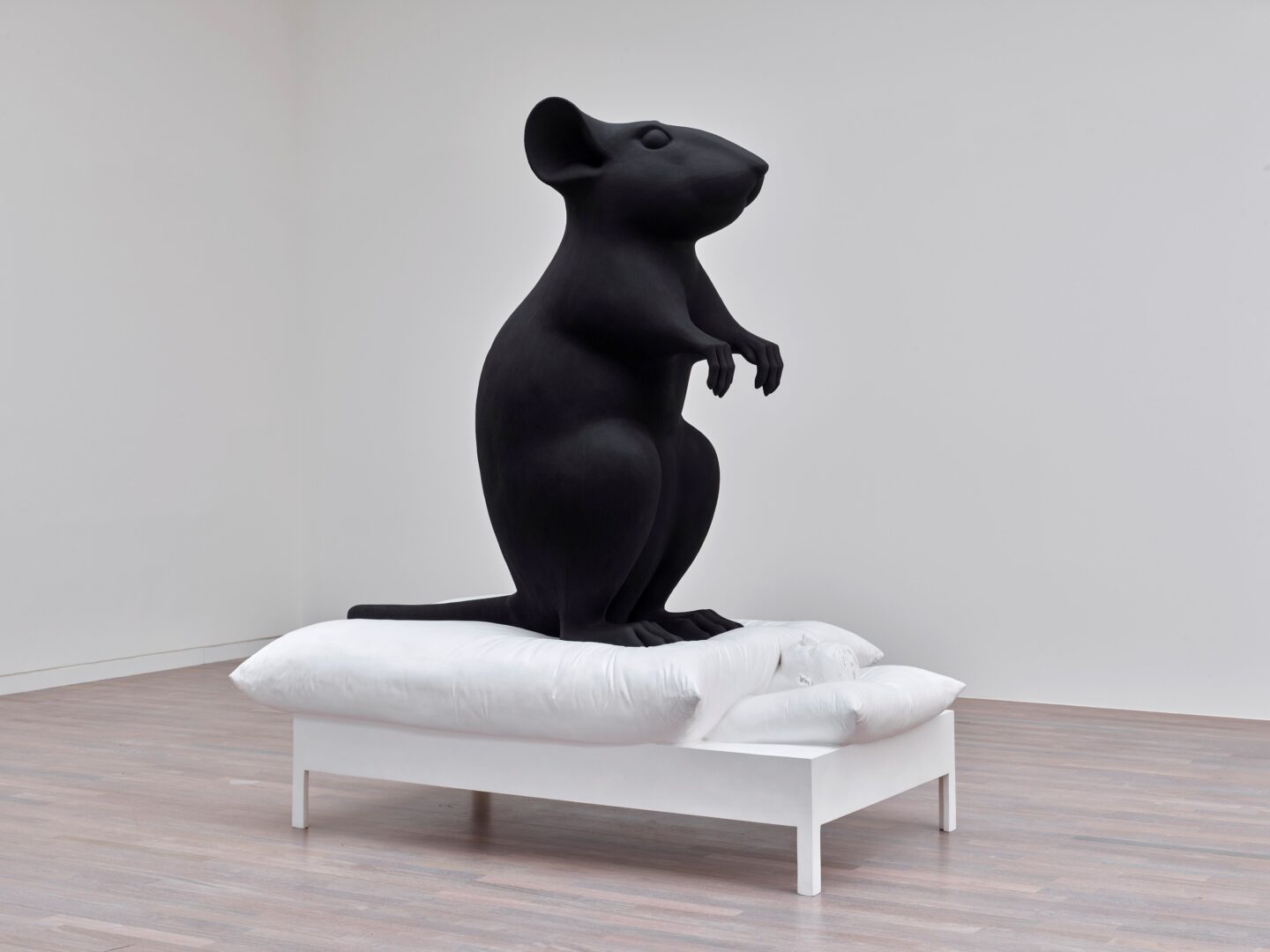 Man sieht die riesige Skulptur einer schwarzen Maus auf einem klein wirkenden weißen Bett. Die Arbeit ist von Katharina Fritsche.