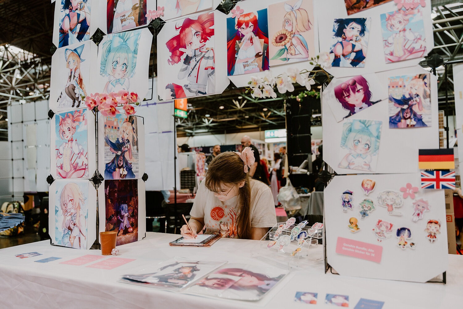 Stand auf der Dokomi, man sieht einen Stand, der rundherum mit Manga-Zeichnungen bestückt ist. In der Mitte ist der Stand offen und man sieht eine junge Frau die zeichnet. 