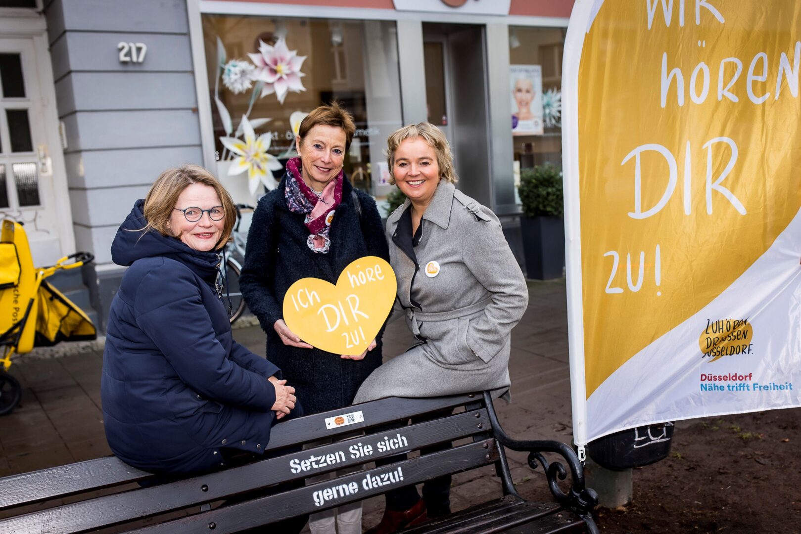 Drei Frauen stehen hinter einer "Zuhör.Bank" in Flingern und halten gemeinsam ein orangfarbenes Herz aus Pappe, auf dem steht: Ich höre dir zu.