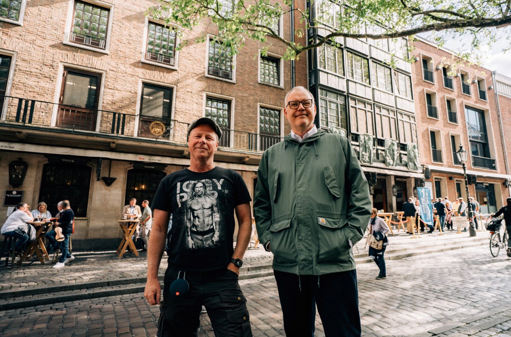 Die Tour-Guides Dirk Mewes (links) und Michael Wenzel stehen an einem sonnigen Tag auf dem Kopfsteinpflaster der Ratinger Straße, im Hintergrund sieht man die alten Gebäude und ihre Gastronomie.