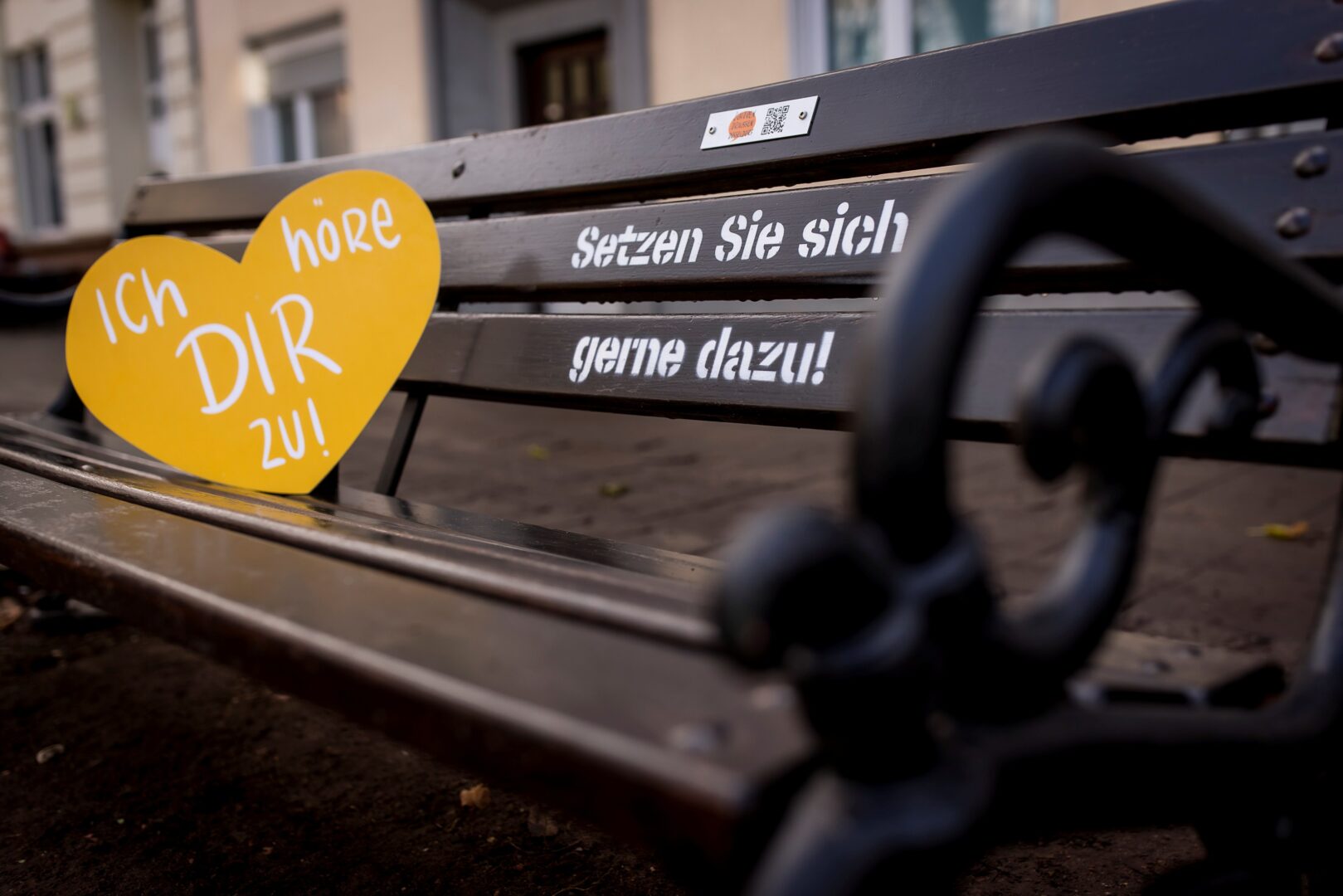 Man sieht eine Bank aus dunklem Holz und an den Seiten mit geschwungenen Lehnen aus Metall. Auf der Bank steht: Setzen Sie sich gerne dazu! Außerdem lehnt ein orangefarbenes Pappherz auf der Sitzfläche mit der Aufschrift: Ich höre dir zu.