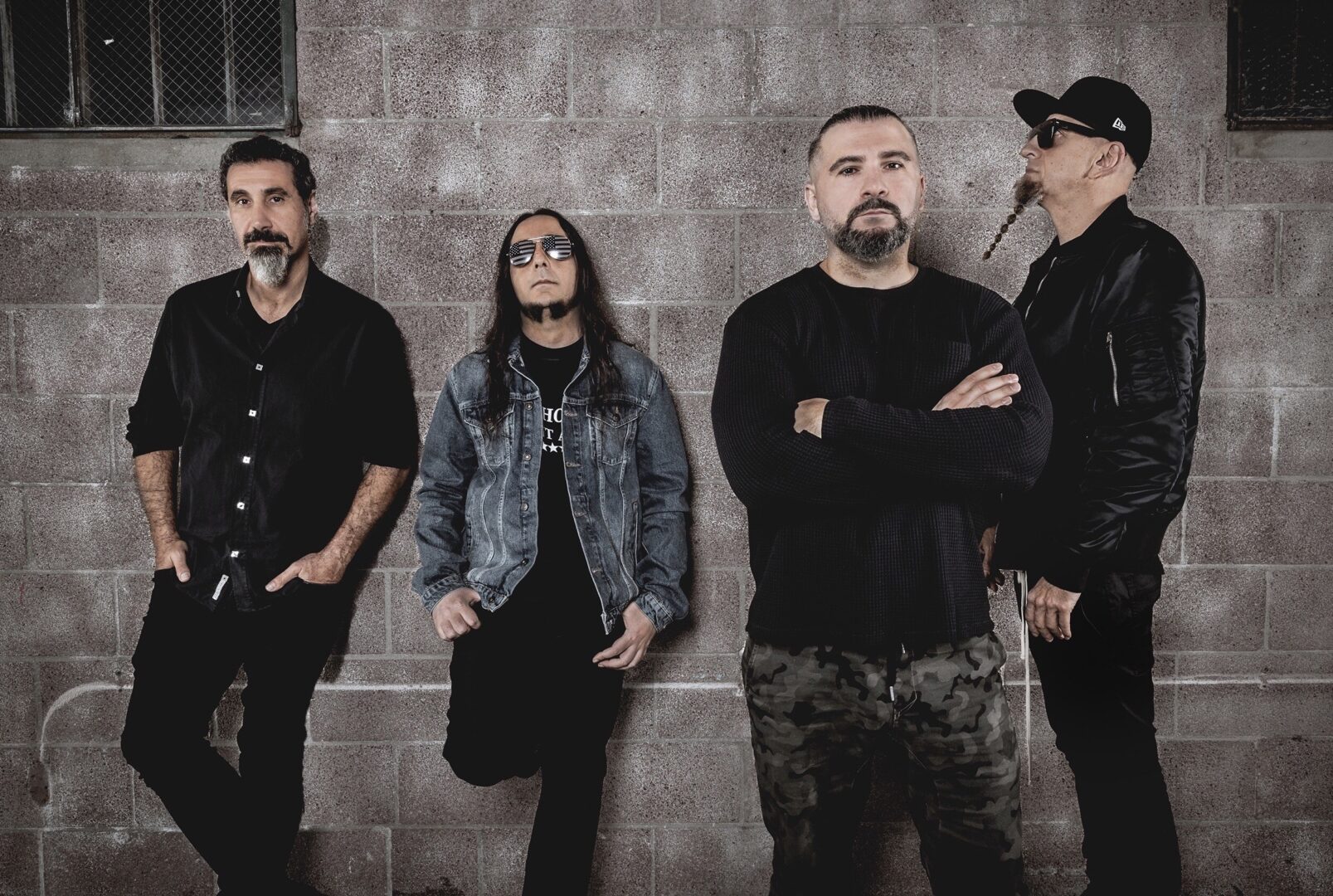 Die Metalband System of a Down. Sie tragen dunkle Bekleidung, gucken ernst und stehen vor einer Wand in verschiedenen Graubrauntönen.