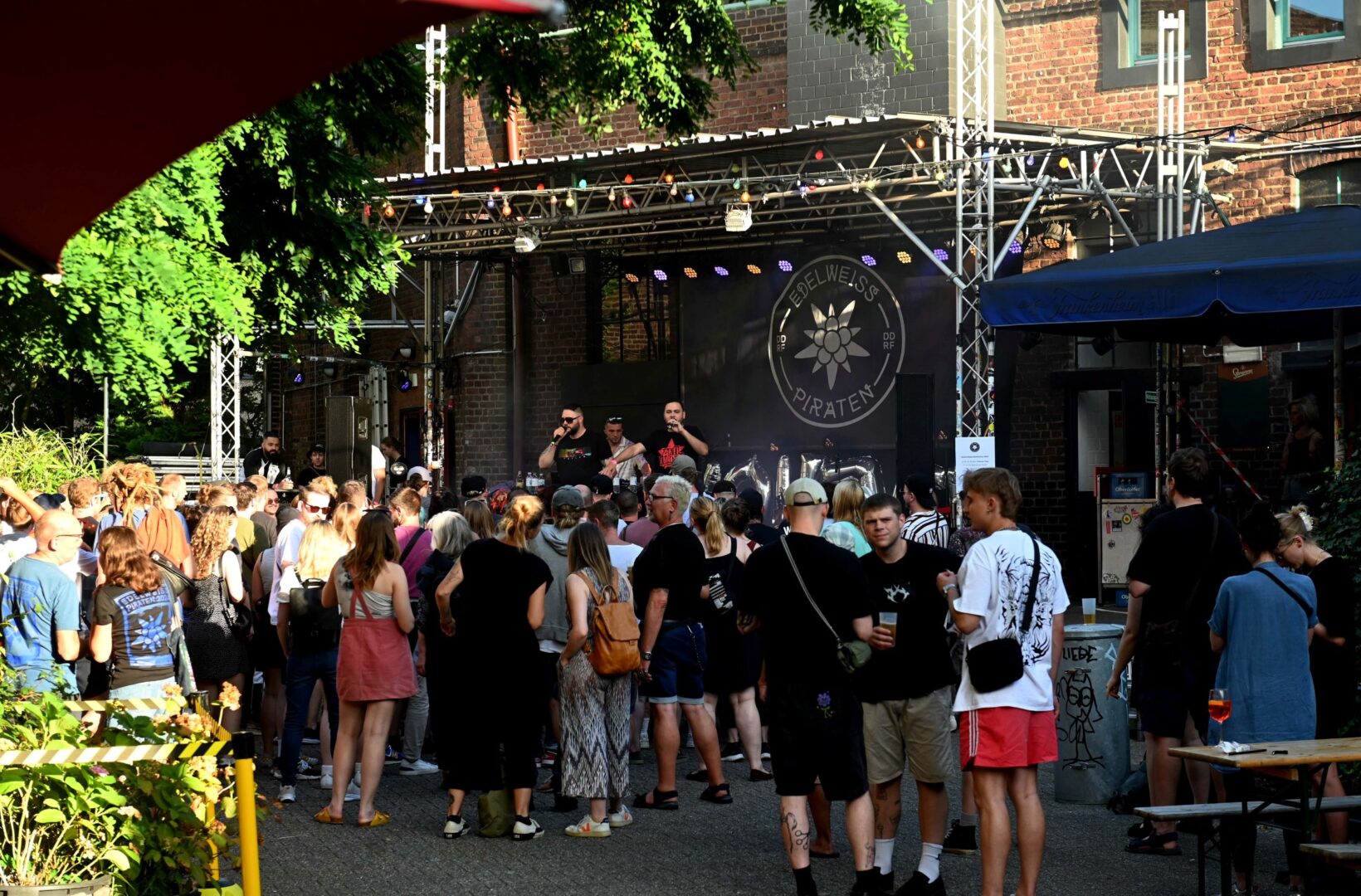 Man sieht den Biergarten des zakk. Es ist Sommer und man sieht eine Bühne mit zwei Künstlern sowie Publikum.