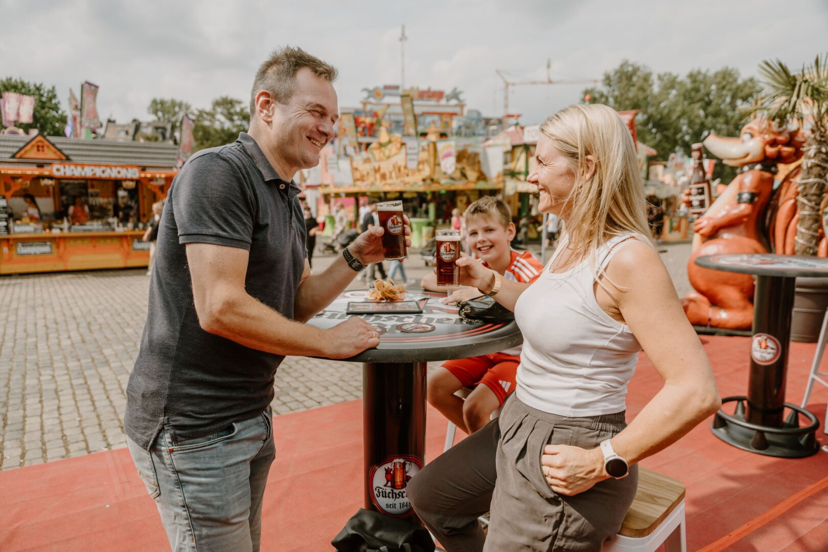 Man sieht ein Ehepaar am Stehttisch und im Hintergrund einen Jungen am Füchschen Bierzelt draußen.