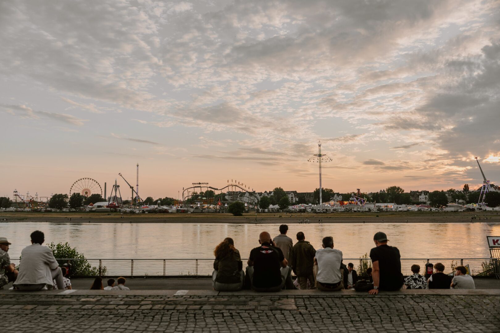 Man sieht Menschen am Rheinufer in der Altstadt von hinten, die über den Rhein auf die Kirmes blicken. Es ist Abend, etwas dunkel, der Himmel changiert von Apricot, blau bis Grau.